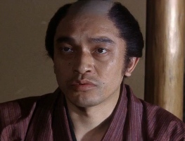 A Japanese man of indeterminate age in period costume, with shaved forehead, gazes at something off-camera