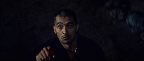 A mature Japanese man in the costume of a priest points upward while talking to someone off-camera