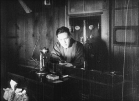 In a production still from a silent movie, a young Japanese man sits in front of an old-style telephone, appearing to be staring at something in his hand